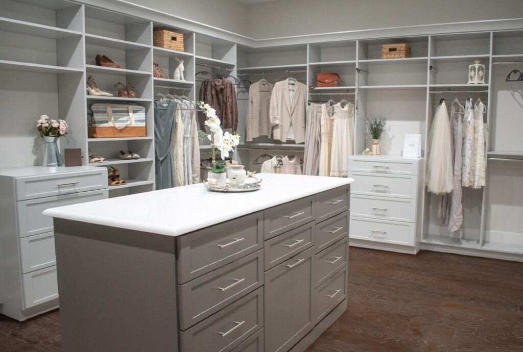 Custom, two-toned closet in light gray with a dark "greige" triple island. It features chrome hardware, crown molding, shoe shelving, multiple drawer stacks, valet rods, and an island filled with drawers and a built-in hamper.