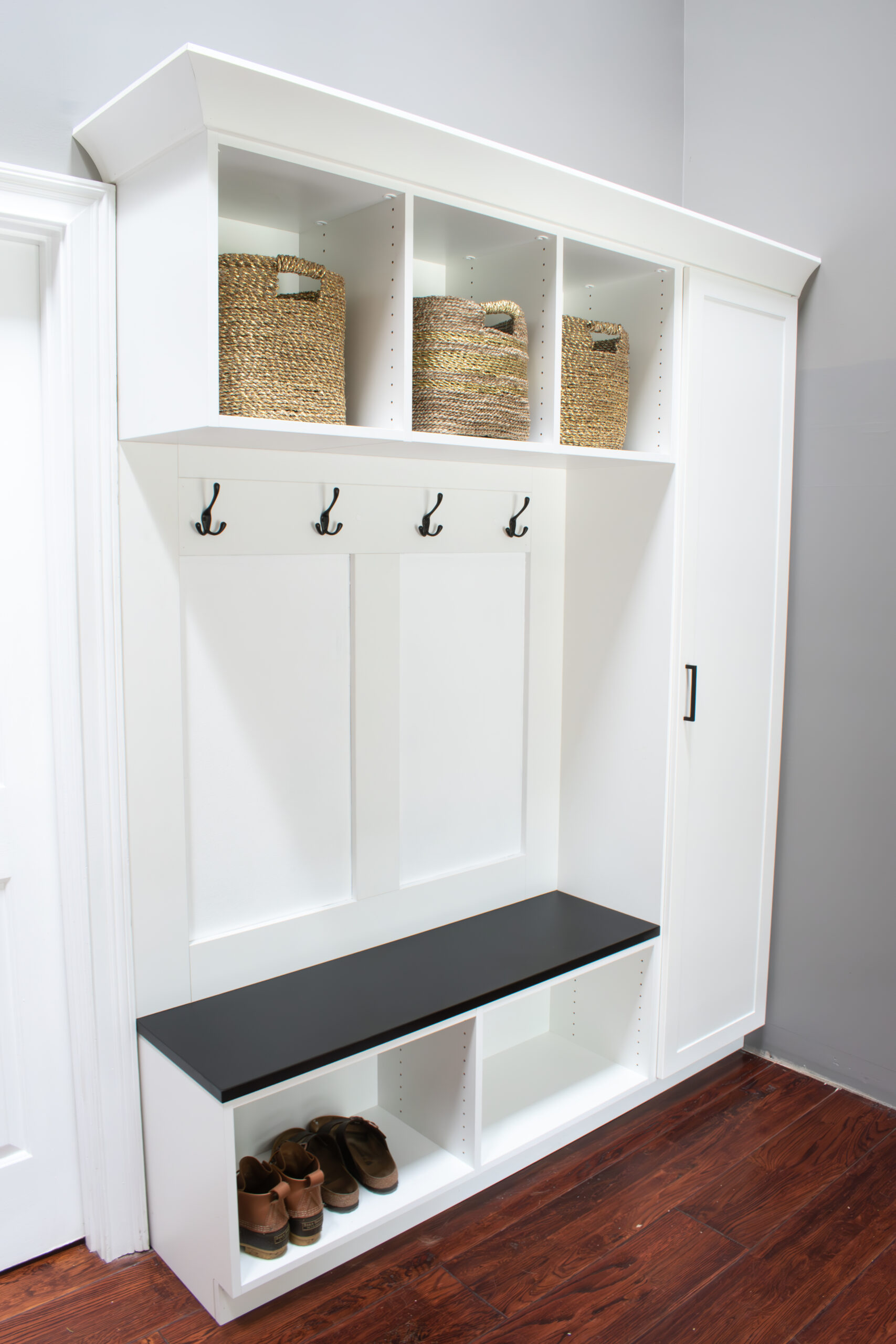 Custom white shaker mudroom with black accents by Custom Closets of GA. It contains one tall cabinet, overhead cubbies, crown molding, coat hooks, a decorative backing, and a bench.
