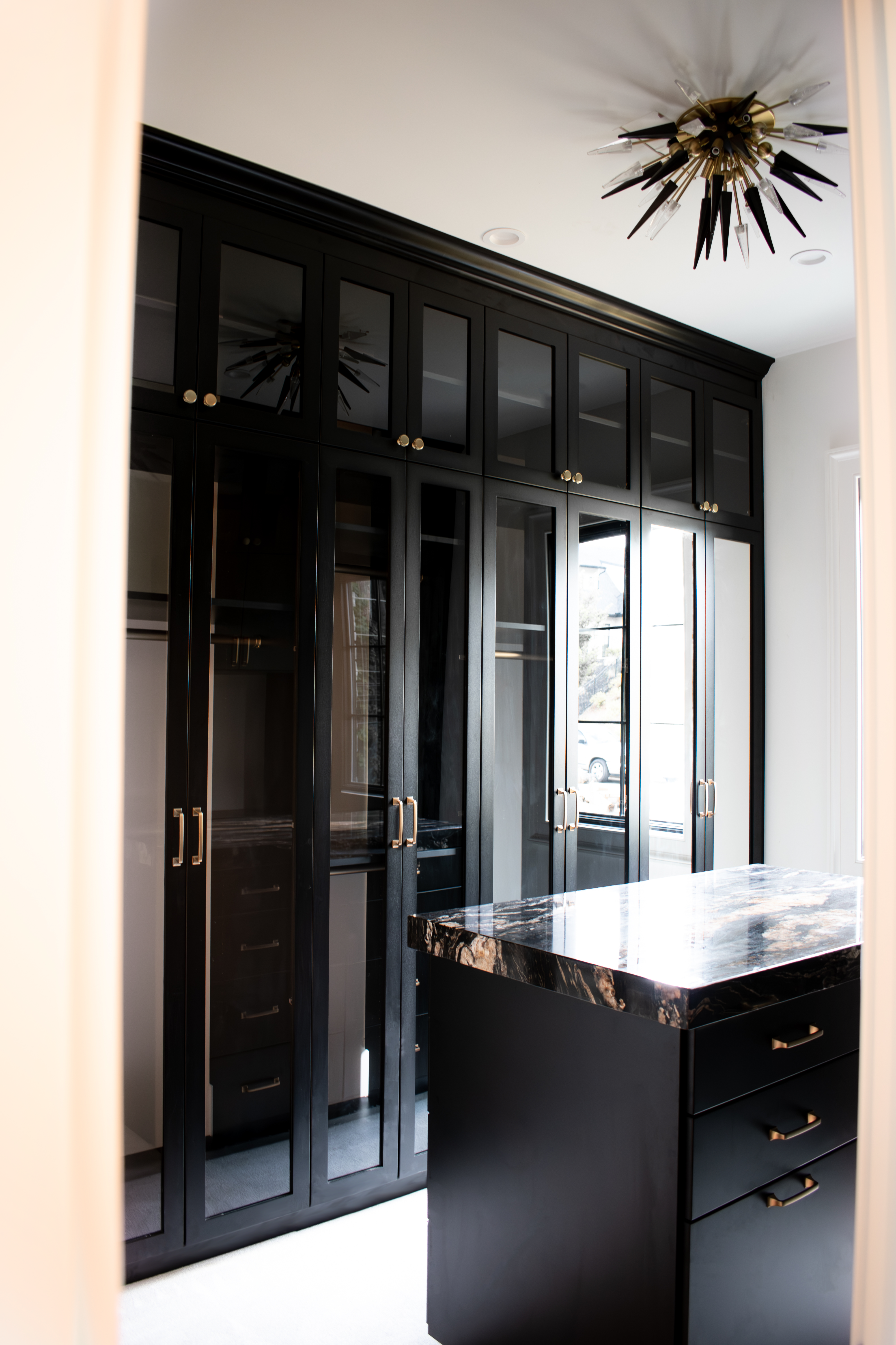 Custom Closets of GA black and gold walk-in closet with island. It contains 24 deep cabinets for hanging clothes, glass and mirrored doors, built-in hampers and drawers, crown molding, and shoe shelves.