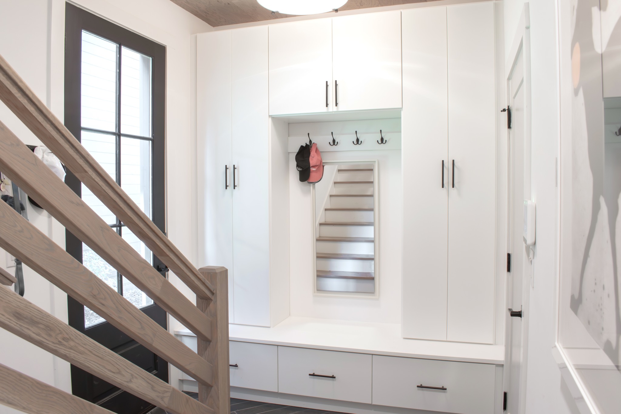 Custom white shaker mudroom with black accents by Custom Closets of GA. It contains one tall cabinet, overhead cubbies, crown molding, coat hooks, a decorative backing, and a bench.